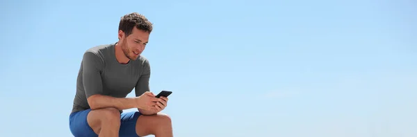 Hombre feliz usando aplicación de teléfono jugando juego en línea sentado fuera después de entrenamiento de gimnasio pancarta panorámica con el fondo del cielo. Fitness atleta estilo de vida activo. —  Fotos de Stock