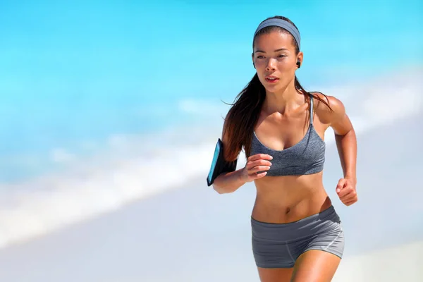 Correr atleta mujer corriendo en la playa escuchando podcast con auriculares inalámbricos y teléfono móvil. Corredor asiático haciendo ejercicio al aire libre estilo de vida activo saludable. — Foto de Stock