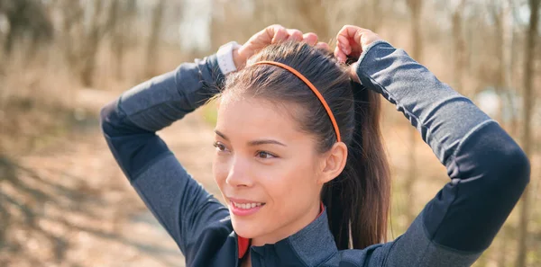 La coureuse asiatique se prépare à courir dehors en attachant ses cheveux dans sa queue de cheval pour un entraînement hiit jogging. Joyeux sourire athlète jogger bannière. — Photo