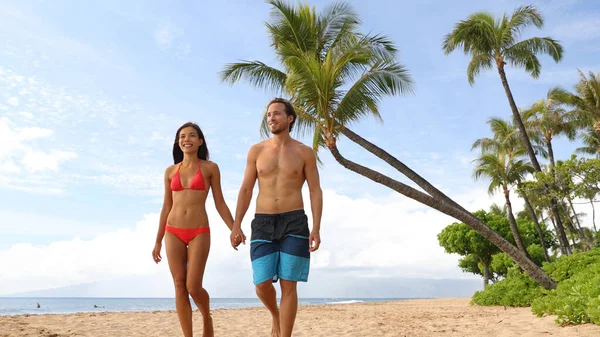 カップルがビーチを歩いている。若い幸せな異人種間のカップルのビーチで水着で手を握って笑みを浮かべて歩く。アジア系女性白人男性 — ストック写真