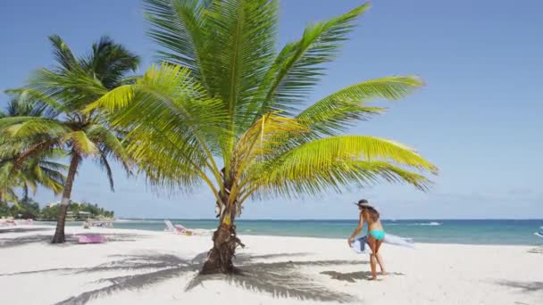 Woman on beach putting down beach towel for sunbathing under palm tree — Stock Video