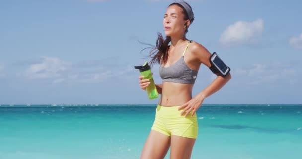 Fáradt atléta futó nő, aki szünetet tart futás közben, kimerülten lélegzik a forró nyári napon, hőgutát kap. Ázsiai futó zenét hallgat vezeték nélküli fülhallgató mobiltelefon app. — Stock videók