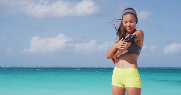 Ouvir podcast no aplicativo de telefone com fones de ouvido sem fio durante o treino na praia. Apto asiático corredor mulher feliz usando wearable tecnologia braçadeira fora — Vídeo de Stock