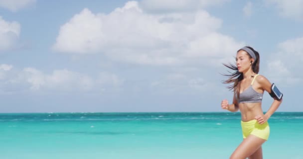 Correr atleta mujer corriendo en la playa escuchando podcast con auriculares inalámbricos y teléfono móvil. Corredor asiático haciendo ejercicio al aire libre estilo de vida activo saludable — Vídeo de stock
