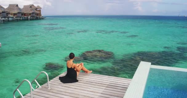 Vacaciones de lujo en Bora Bora. Elegante mujer en vestido negro relajante disfrutando de la vista de los arrecifes de coral en el océano Polinesia francesa en el hotel resort bungalow sobre el agua — Vídeos de Stock