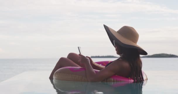 Conexão rápida do Internet do telefone 5G na mulher das férias no flutuador inflável do donut usando o telefone celular na piscina. Menina relaxante relaxante desfrutando de férias de viagem na piscina resort em biquíni. — Vídeo de Stock