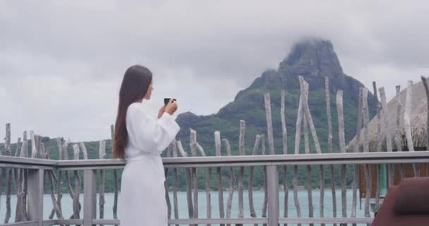 Γυναίκα πίνοντας πρωινό καφέ χαλαρώνοντας φορώντας μπουρνούζι κοιτάζοντας τη θέα του ωκεανού και του όρους Otemanu στο Bora Bora, Γαλλική Πολυνησία, Ταϊτή. Πολυτελείς διακοπές. Ασιάτης επισκέπτης που απολαμβάνει θέα — Αρχείο Βίντεο