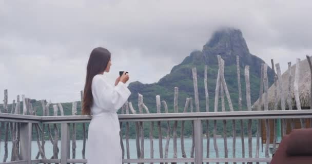 Mujer tomando café de la mañana relajante con albornoz mirando al océano y el Monte Otemanu en Bora Bora, Polinesia Francesa, Tahití. Vacaciones de lujo. Asiático viaje huésped disfrutando vista — Vídeo de stock