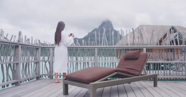 Travel vacation luxury. Woman drinking morning coffee relaxing wearing bathrobe looking at view of ocean and mountain landscape. Luxury living resort lifestyle at Bora Bora, French Polynesia, Tahiti. — Stock Video