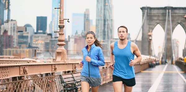 Futó pár futók kocognak a város utcáján az eső zászlaja alatt. Ázsiai nő és kaukázusi férfi futó és fitness sport modellek edzés maraton szabadban a Brooklyn Bridge, New York City — Stock Fotó