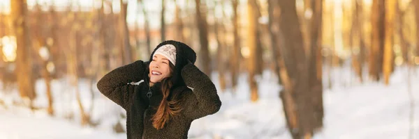 Hiver froid femme par temps protéger les oreilles tenant chapeau chaud sur cache-oreilles forêt fond bannière panorama. Frostbite protection vêtements de dessus asiatique fille dans la neige portant manteau veste en peluche sherpa, gants — Photo