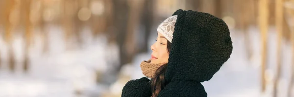 Invierno mujer feliz relajante respirar aire frío fuera de los jóvenes estilo de vida. Chica asiática con los ojos cerrados respirando bienestar y salud en el bosque con chaqueta encapuchada y sombrero bandera panorámica — Foto de Stock