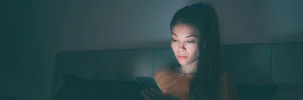 Yatakta cep telefonu kullanan Asyalı kadın uykusuzluk çekiyor. Üzgün kız cep telefonundan sosyal medyaya bakıyor. Koyu panoramik pankartta parlayan mavi ışık — Stok fotoğraf