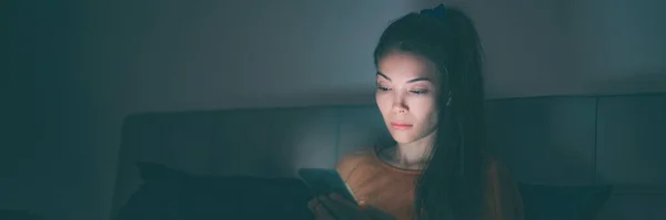 Telefoon in bed Aziatische vrouw met behulp van mobiele telefoon late avond slapeloosheid - triest meisje depressief kijken naar sociale media op mobiele telefoon. Blauw licht gloeiend in de donkere panoramische banner — Stockfoto