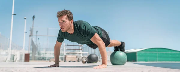 Fitness Man πανόραμα πανόραμα. Αθλητική δύναμη προπόνηση pushup εξισορρόπηση πόδια για μπάλα ιατρικής για προχωρημένο σώμα προπόνηση push-ups πάτωμα ασκήσεις σε υπαίθριο γυμναστήριο — Φωτογραφία Αρχείου
