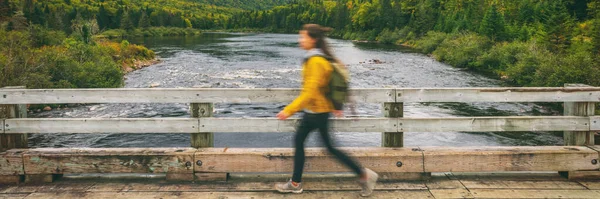 등산객 여자가 배낭을 메고 강을 건너 다리 위를 걷고 있어요. 야외 자연 풍경을 여행하는 관광객들의 움직임은 가을 배경의 파노라마 현수막을 타고 내려온다. 퀘벡, 캐나다 — 스톡 사진