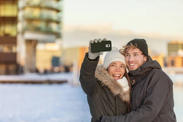 Selfie para szczęśliwy spacer zimowy turyści biorąc zdjęcie z telefonu na ulicy miasta panorama stylu życia. Azjatka, biały mężczyzna w dzianinach czapki i kurtki — Zdjęcie stockowe