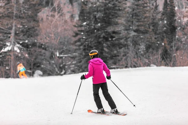 Sciatrice che impara a sciare sulle piste di montagna. Sport invernali attività all'aria aperta. Ragazza da dietro in giacca rosa e occhiali, casco, sci, bastoncini — Foto Stock
