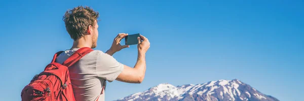Travel tourist hiking man taking picture with phone of mountains landscape on summer hike adventure. Panorama banner with copy space on blue sky background — Stock Photo, Image