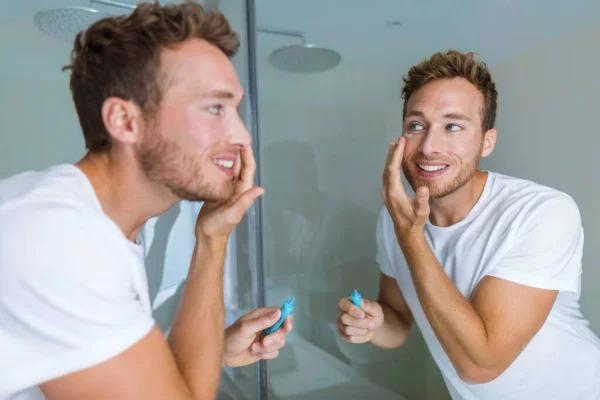 Man putting skincare facial treatment cream on face. Anti-aging skin care product. Male beauty morning routine at home lifestyle. Guy looking in bathroom mirror applying moisturizer under eyes — Stock Photo, Image