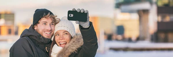 Selfie çifti mutlu kış yürüyüşleri turistleri şehir sokaklarındaki panorama yaşam tarzında telefonla fotoğraf çekiyor. Asyalı kadın, beyaz adam örgü şapka ve ceket giyiyor. — Stok fotoğraf