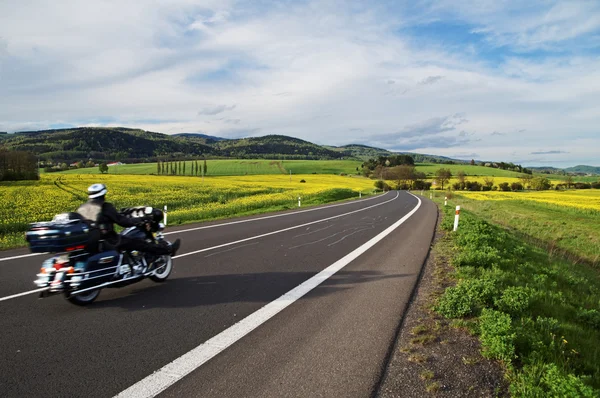 Motorcycle traveling along an empty asphalt road between yellow blooming rape fields — Stock Photo, Image