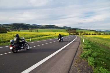 Motorcycles traveling along an empty asphalt road between yellow blooming rape fields clipart
