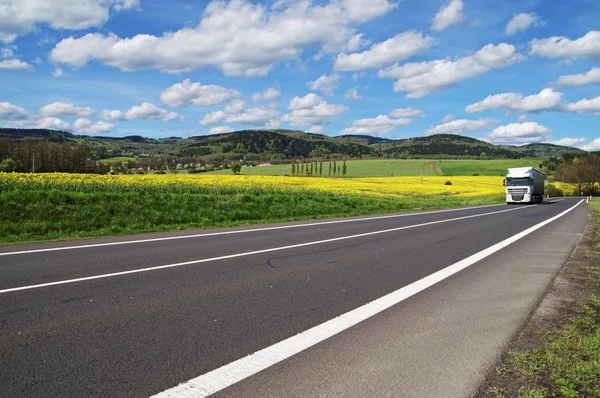 Camion bianco arriva da lontano su una strada asfaltata tra il campo di colza fiorito giallo — Foto Stock