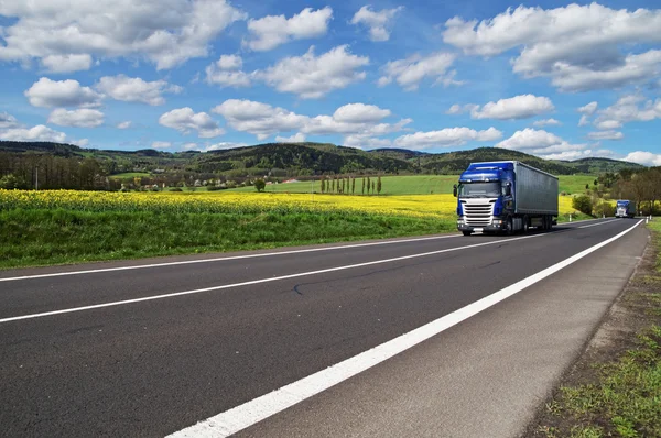 Two blue trucks driving on asphalt road between yellow flowering rapeseed