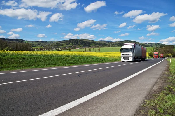 Lastbilar som kör vägen runt fältet gula blommande raps i landsbygdens landskap — Stockfoto