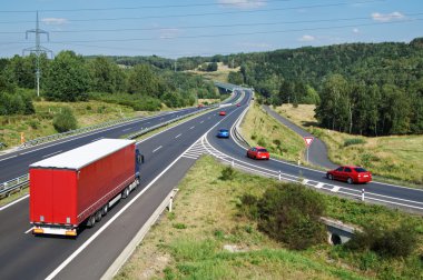 Asfalt karayolu Kırmızı kamyon ve kırmızı binek otomobilleri ormanlık ülke ile. Slip yol trafik işaretleri ile yol vermek. Geçis kapı uzakta. Yukarıdan görüntüleyin. Mavi gökyüzü gün güneşli yaz.