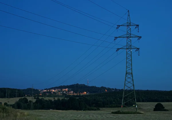 High power line pylons are intended to electrically lighted city in the background. Rural landscape in the evening. — Stock Photo, Image