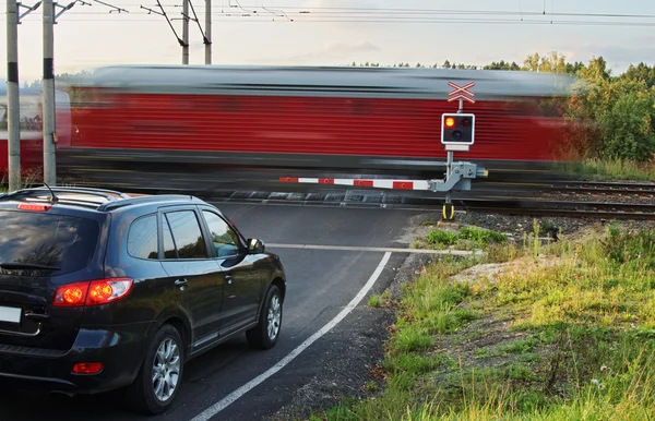 Speeding motion blur red train passing through a railway crossing with gates. Black car standing in front of the railway barriers on an asphalt road. — Stock Photo, Image