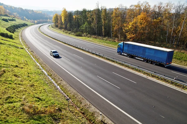 Dálnice mezi opadavé lesy s listy v barvách podzimu, dálnice pokračuje modrá kamionu a osobního automobilu — Stock fotografie