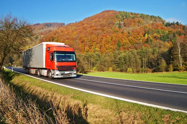 Rode vrachtwagen op de weg onder de beboste berg van kleurrijke herfst kleuren Stockfoto