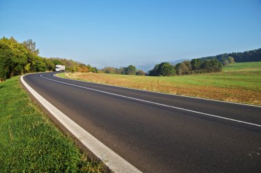 Asphalt road in the countryside, white truck coming around in the distance the bend clipart