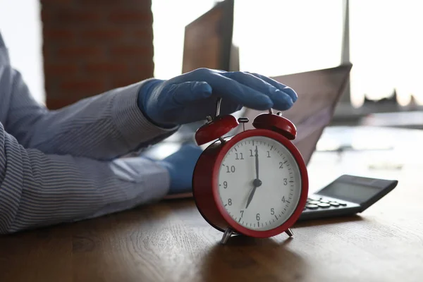 Žena s modrými ochrannými rukavicemi držet červené budík 7 hodin proti modernímu kancelářskému stolu detailní — Stock fotografie