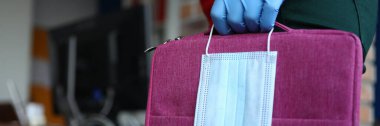 Güvenlik eldivenli bir kadın maske ve laptop çantasını yakın çekime karşı tutuyor.