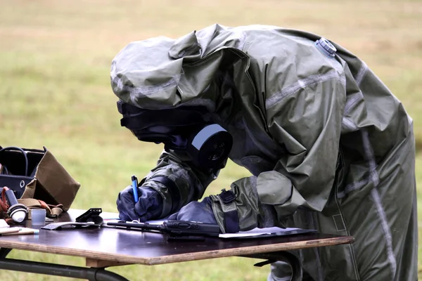 Man in gas mask and special safe suit for biological danger writing notes on the table. Field on background. Biological or nuclear or gas danger.