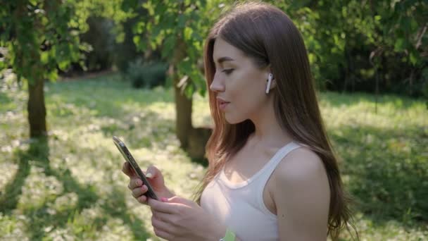 Ritratto di giovane bella donna che ascolta musica in auricolare senza fili dal suo telefono sullo sfondo di alberi verdi estivi nel parco — Video Stock