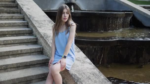 Mooie jonge vrouw zit bij fontein in blauwe jurk poseren voor de camera en wachten op iemand. slow motion — Stockvideo