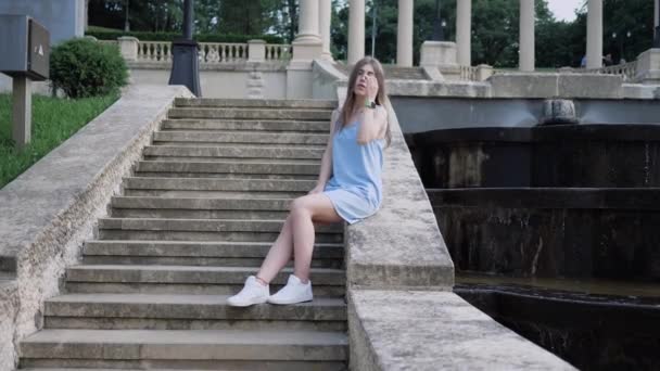 Красивая молодая модель в голубом платье сидит на лестнице возле каскадного фонтана и позирует перед камерой в жаркий летний день. замедленное движение — стоковое видео