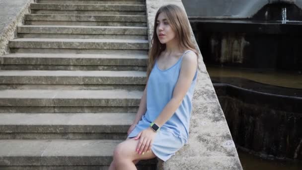 Eine junge hübsche Frau in blauem Kleid sitzt auf einer Treppe und ruht sich an einem heißen Sommertag aus. Zeitlupe — Stockvideo