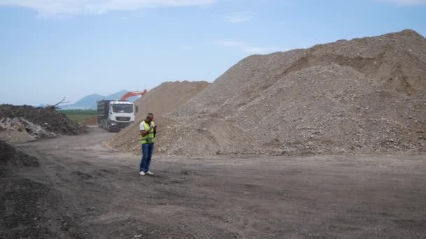 Foreman em canteiro de obras com câmera em suas mãos, usando telefone celular. Contra o fundo de grandes montanhas de escombros e equipamentos de construção. Rússia, Stavropol, 10.06.20 — Vídeo de Stock