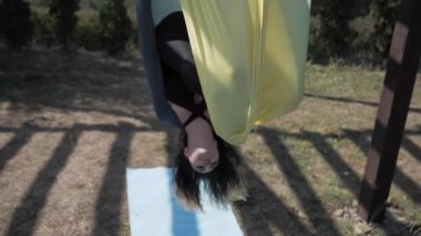 Park 'taki hamakta hava yogası yapan atletik genç bir kadın karmaşık akrobatik egzersizler yapıyor.