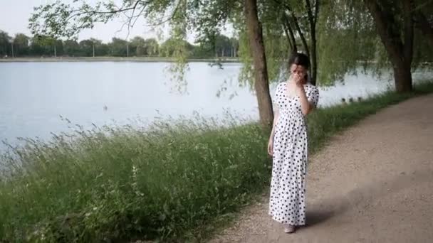 Junge schöne Frau im weißen langen Kleid mit langen Haaren spaziert am See entlang, Park — Stockvideo