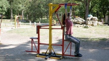 Eldivenli ve maske takan genç bir kadın Park 'taki spor aletlerinde. Halka açık yerlerde simülatör kullanımında koronavirüs salgınından korunma.