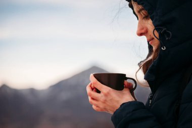Genç bir kadın, elinde bir bardak sıcak içkiyi dağların arka planında tutuyor..