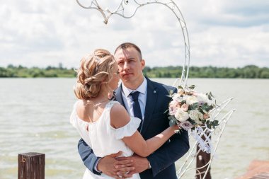 Güzel gelin ve damat bohem tarzında bir elbiseyle yazın göl kenarındaki bir rıhtımda kalp şeklinde çelik kuğulara karşı dururlar.