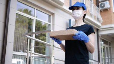 Postacı, koruyucu maskeli kurye ve tıbbi eldiven taşıyan küçük bir paket taşıyor. Karantina altında teslimat servisi, salgın hastalıklar. Sosyal uzaklık kavramı.